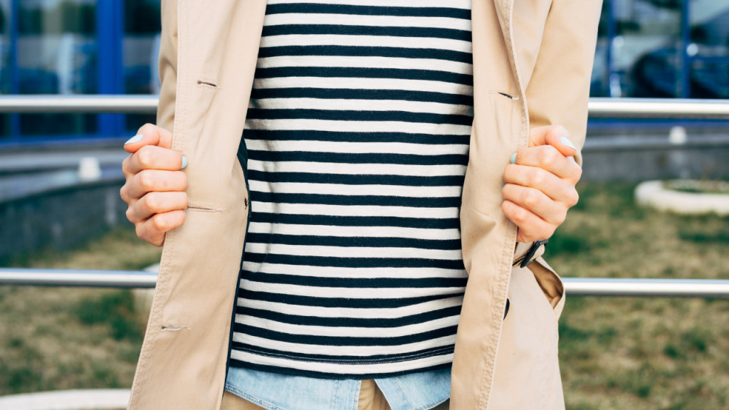 Person wearing a beige coat over a black and white striped shirt, standing outdoors and holding the coat open with both hands, showcasing a chic french style.